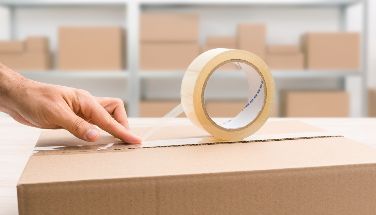Roll of clear hot melt carton sealing tape being applied to seal a cardboard shipping box in a warehouse